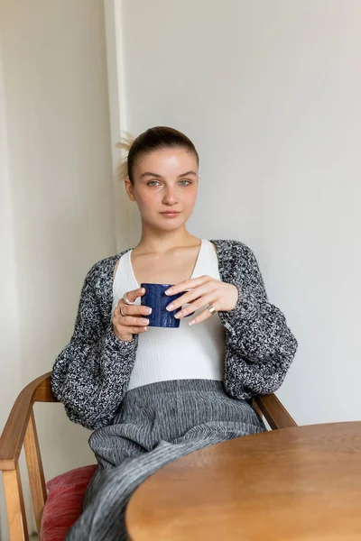 Woman in warm cardigan holding cup and looking at camera at home — Stock Photo