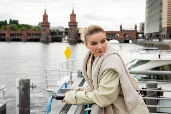Jeune femme en trench coat tenant un sac d'embrayage sur la jetée avec pont Oberbaum à l'arrière-plan à Berlin — Photo de stock