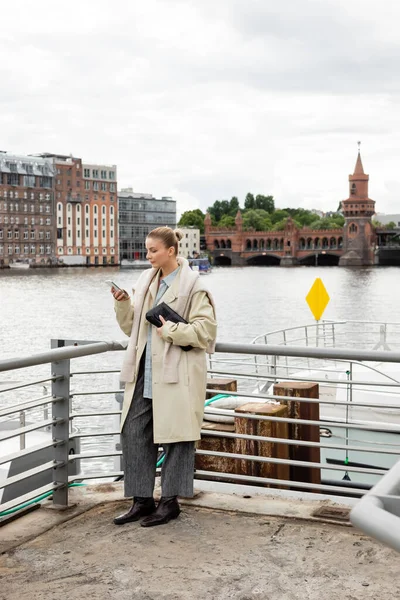 Mulher de casaco de trincheira usando smartphone e segurando saco de embreagem no cais em Berlim — Fotografia de Stock