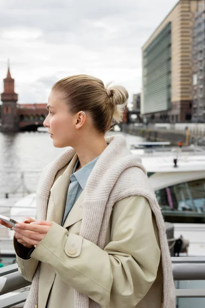 Бічний вид на жінку в плащі, що тримає смартфон на міській вулиці в Берліні. — стокове фото