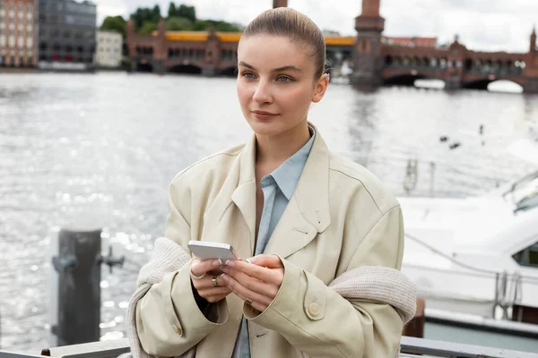 Junge Frau im Trenchcoat hält Smartphone in der Hand und schaut auf Straße in Berlin weg — Stockfoto