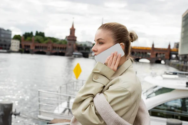 Vista laterale della donna in trench che parla sul cellulare vicino al fiume a Berlino — Foto stock