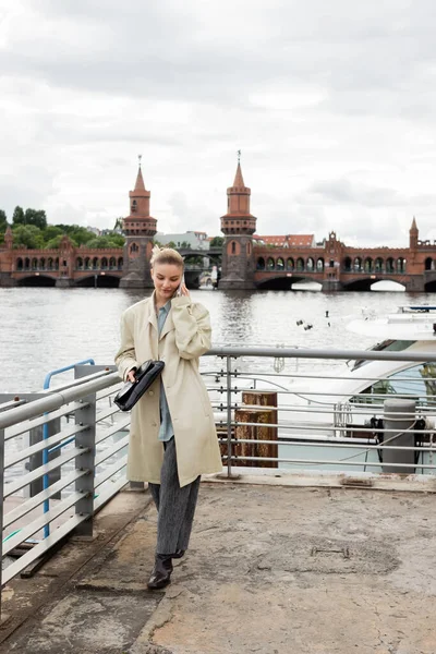 Jeune femme en trench coat tenant un sac d'embrayage et parlant sur smartphone à Berlin — Photo de stock