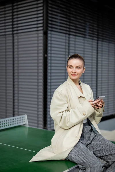 Femme en trench coat à l'aide d'un smartphone assis sur une table de ping-pong dans la rue — Photo de stock