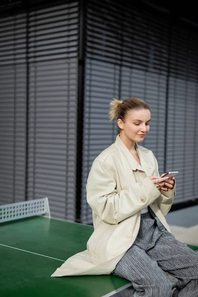 Young woman in trench coat holding smartphone while sitting on ping-pong table outdoors — Stock Photo