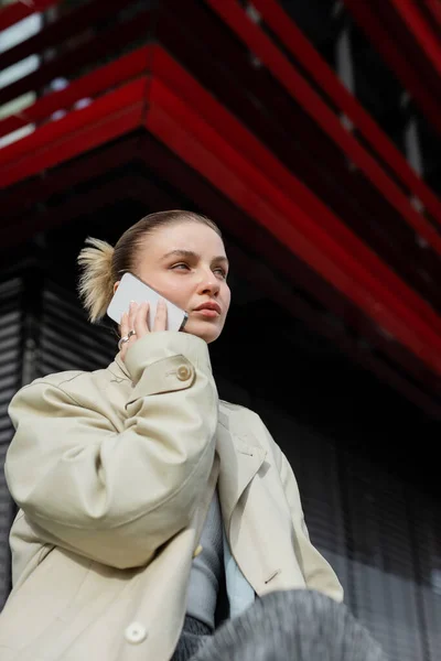 Низкий угол обзора женщины в плаще разговаривающей на улице со смартфоном — стоковое фото