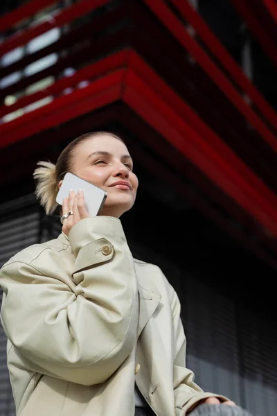 Low angle view of smiling woman in trench coat talking on smartphone outdoors — Stock Photo