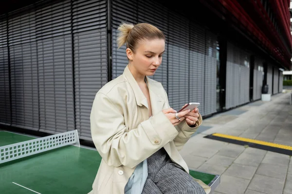 Donna in trench utilizzando il cellulare vicino a ping pong sulla strada urbana — Foto stock