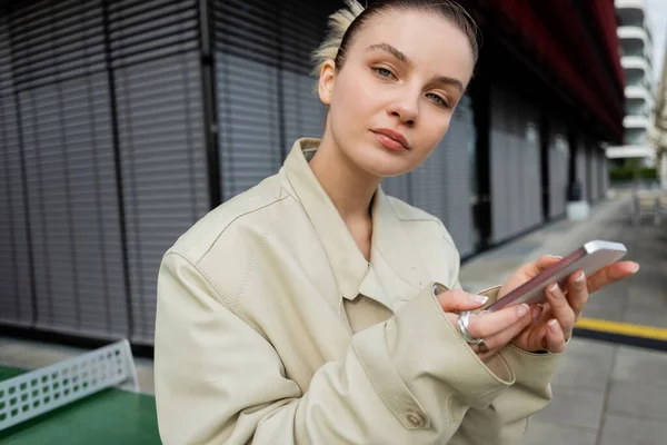 Mujer joven en gabardina sosteniendo teléfono inteligente y mirando a la cámara en la calle urbana - foto de stock