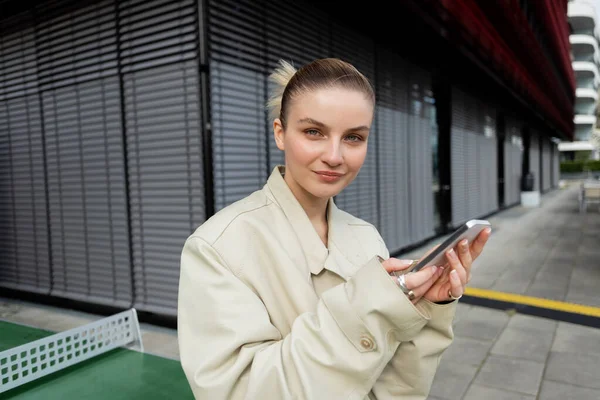Mujer sonriente en gabardina sosteniendo teléfono inteligente y mirando a la cámara en la calle urbana - foto de stock