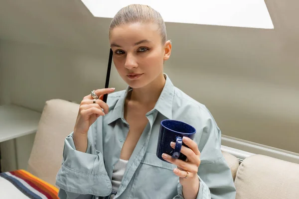Junge Frau hält Stift und Tasse in der Hand, während sie auf der Couch im Wohnzimmer in die Kamera blickt — Stockfoto