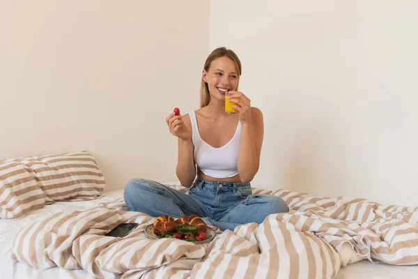 Donna sorridente che tiene pomodoro ciliegia e succo d'arancia vicino a cornetti sul letto — Foto stock