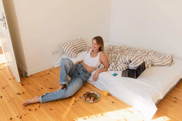 Positive woman sitting near laptop and breakfast in bedroom — Stock Photo