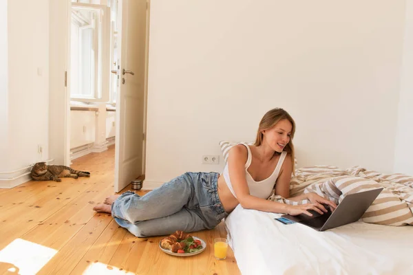 Mujer sonriente utilizando el ordenador portátil cerca de la tarjeta de crédito y desayuno en el dormitorio - foto de stock