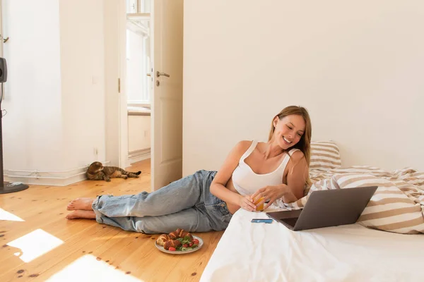 Junge Frau hält Orangensaft in der Nähe von Laptop und Frühstück im Schlafzimmer — Stockfoto