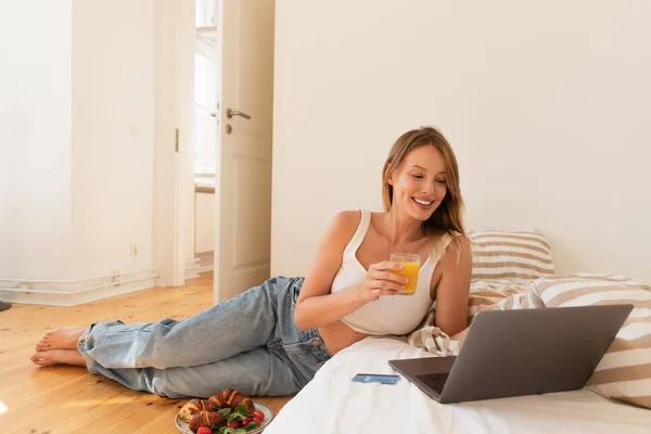 Jovem alegre segurando suco de laranja perto do laptop e café da manhã no quarto — Fotografia de Stock