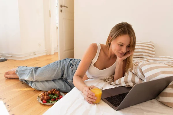 Freelancer sonriente mirando a la computadora portátil y sosteniendo jugo de naranja cerca de sabroso desayuno en el suelo en casa - foto de stock