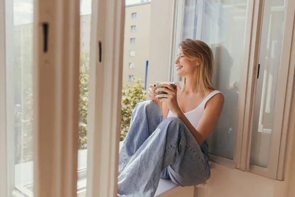 Donna sorridente in jeans che tiene la tazza di caffè mentre si siede sul davanzale della finestra vicino alla finestra aperta a casa — Foto stock