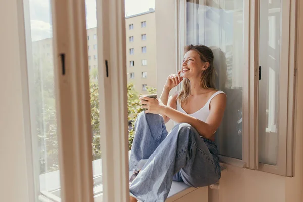 Cheerful young woman holding cup while sitting on windowsill at home — Stock Photo