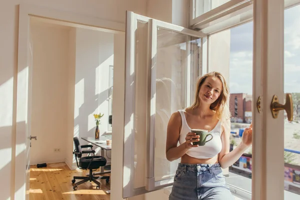 Улыбающаяся женщина в верхней держа чашку возле открытого окна дома — стоковое фото