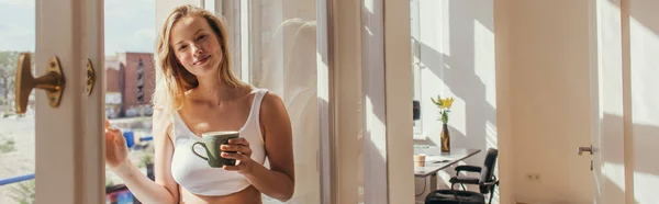 Femme blonde en haut tenant une tasse de café près de la fenêtre ouverte à la maison, bannière — Photo de stock