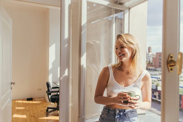 Cheerful young woman in top holding cup near open window at home — Stock Photo