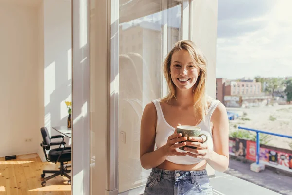 Happy blonde woman in top holding cup and looking at camera near open window at home — Stock Photo