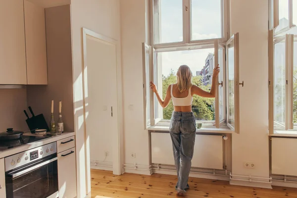 Vue arrière de la jeune femme ouvrant la fenêtre près de la tasse sur le rebord de la fenêtre à la maison — Photo de stock