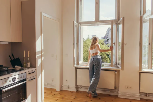 Femme blonde regardant la fenêtre ouverte près de la tasse de café sur le rebord de la fenêtre dans la cuisine — Photo de stock