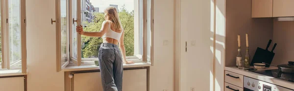 Mujer joven mirando a la ventana abierta cerca de la taza en el alféizar de la ventana en la cocina, pancarta - foto de stock