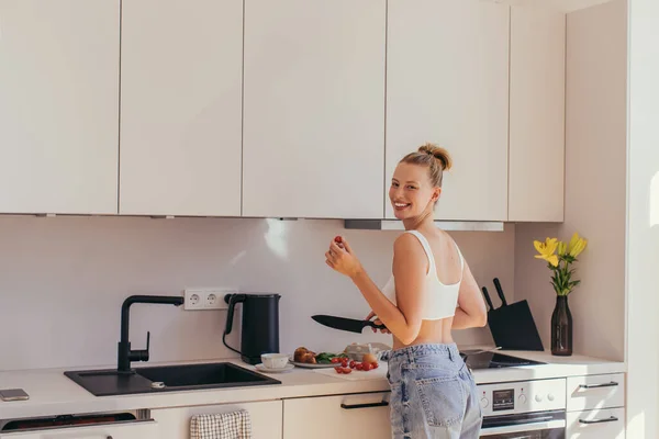 Cheerful young woman in top holding cherry tomato while cooking breakfast in kitchen — Stock Photo