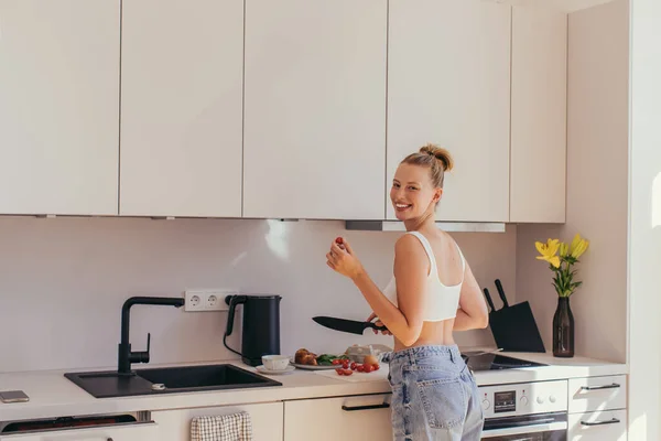 Jovem alegre no topo segurando tomate cereja enquanto cozinha café da manhã na cozinha — Fotografia de Stock