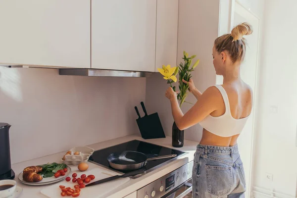 Mulher loira colocando flores em vaso perto do café da manhã e café na cozinha — Fotografia de Stock