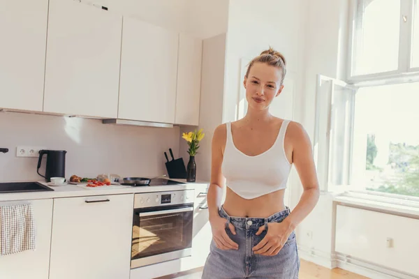 Giovane donna in alto e jeans guardando la fotocamera in cucina — Foto stock