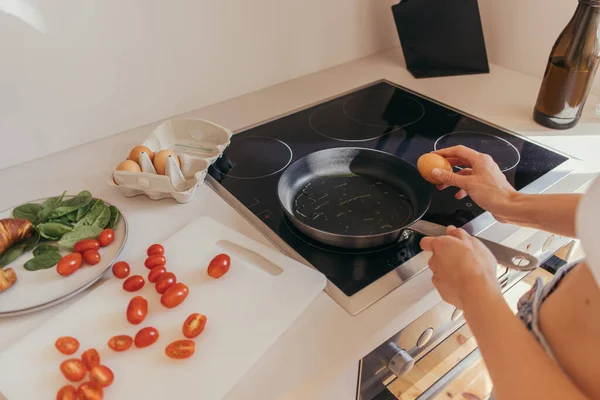 Vista recortada de la mujer sosteniendo huevo cerca de sartén y tomates cherry en la cocina - foto de stock