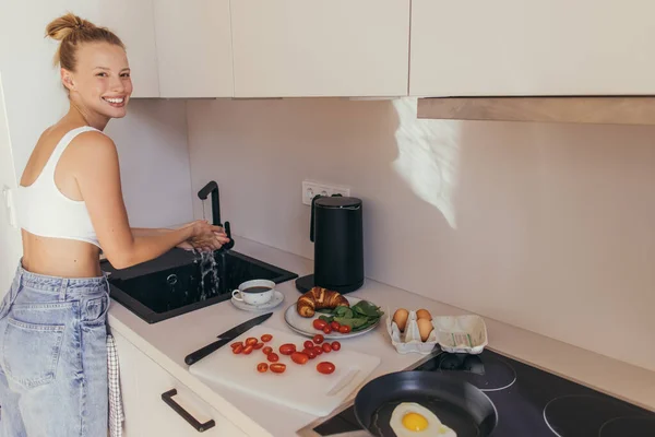 Donna sorridente che guarda la fotocamera e si lava le mani mentre prepara la colazione in cucina — Foto stock
