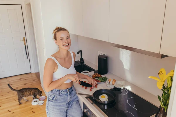 Donna felice guardando la fotocamera mentre taglia pomodorini ciliegia vicino all'uovo sulla padella in cucina — Foto stock