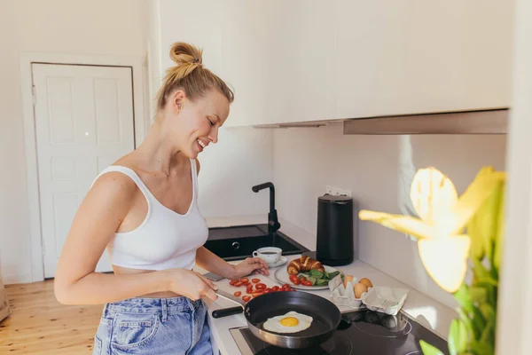 Vista laterale della donna bionda sorridente che guarda l'uovo sulla padella vicino al caffè e ai pomodorini in cucina — Foto stock