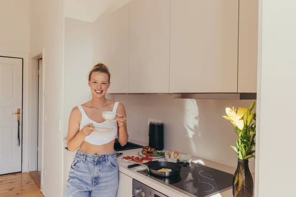 Donna sorridente in cima che tiene la tazza di caffè vicino a colazione e fiori in cucina — Foto stock