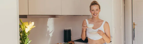 Mujer positiva sosteniendo taza de café cerca de flores de lirio en la cocina, pancarta - foto de stock