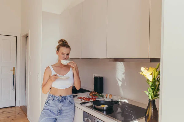 Young woman in top holding coffee cup near breakfast and lily flowers in kitchen — Stock Photo