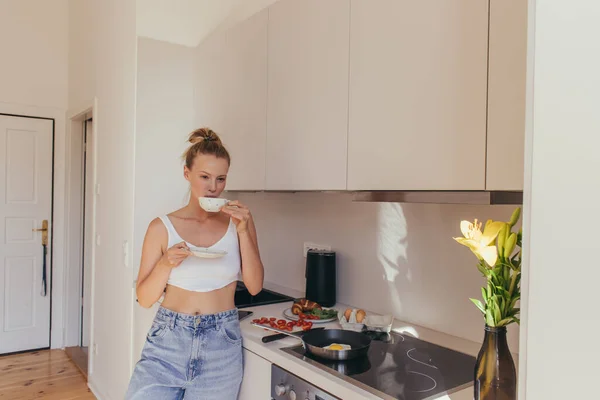 Mujer joven en la parte superior sosteniendo taza de café cerca del desayuno y flores de lirio en la cocina - foto de stock