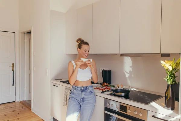 Souriant jeune femme tenant une tasse de café près du petit déjeuner dans la cuisine — Photo de stock