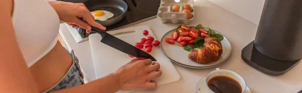 Vista recortada de la mujer cortando tomates cherry cerca de café y croissant en la cocina, pancarta - foto de stock