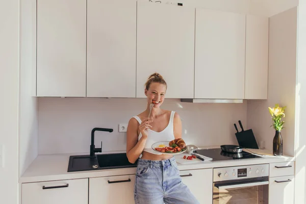 Sorrindo mulher comendo tomate cereja enquanto segurando café da manhã na cozinha — Fotografia de Stock