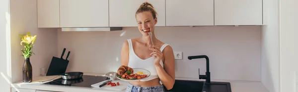 Femme blonde positive mangeant de la tomate cerise et tenant le petit déjeuner dans la cuisine, bannière — Photo de stock