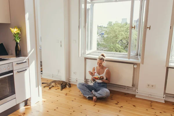 Mujer rubia sonriente sosteniendo plato con desayuno cerca de gato plegable escocés en el suelo en la cocina - foto de stock