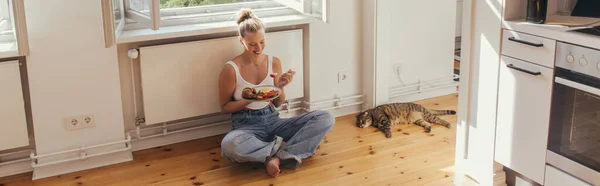 Donna allegra che tiene il piatto con deliziosa colazione vicino al gatto pieghevole scozzese sul pavimento a casa, striscione — Foto stock