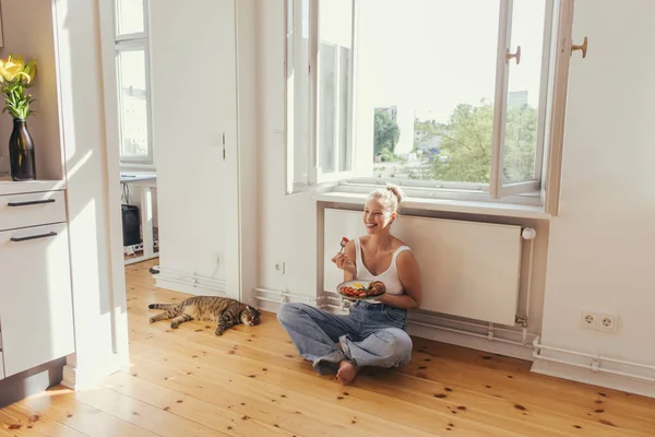Femme souriante en jeans tenant la plaque avec petit déjeuner près du chat pliant écossais sur le sol à la maison — Photo de stock