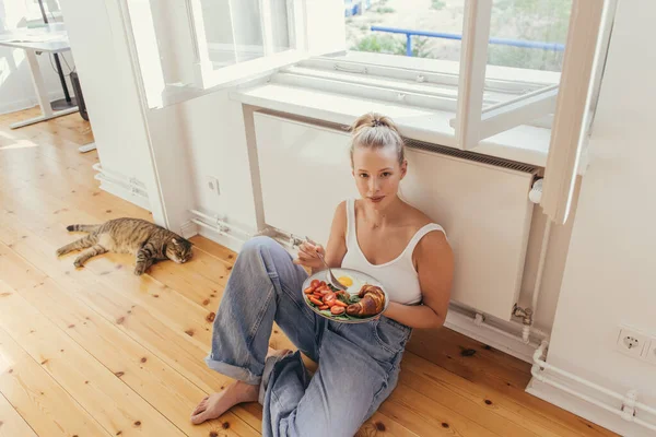 Hochwinkelaufnahme der blonden Frau mit Teller und Frühstück und Blick in die Kamera in der Nähe der schottischen Faltkatze zu Hause — Stockfoto